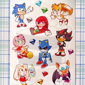 May include: A sticker sheet featuring characters from the Sonic the Hedgehog video game series. The sheet includes Sonic, Tails, Knuckles, Shadow, Metal Sonic, Amy Rose, Cream the Rabbit, and Rouge the Bat. The characters are depicted in a cartoon style and are surrounded by colorful gems.