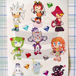 May include: A sticker sheet featuring characters from the Sonic the Hedgehog franchise. The sheet includes Sonic, Tails, Knuckles, Amy, Shadow, Rouge, Blaze, and Cream. The characters are depicted in a chibi style and are surrounded by colorful gems.