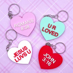 May include: Four heart-shaped keychains in pink, mint, white, and red. Each keychain has a silver ring and chain. The pink keychain reads "ROMANS 8:38", the mint one says "UR LOVED", the white one says "JESUS LUV U", and the red one says "JOHN 3:16".