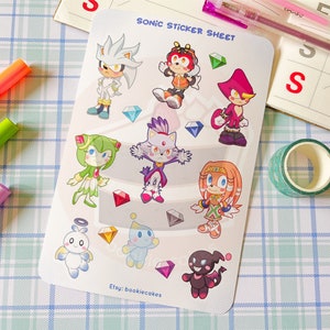 May include: A sticker sheet featuring characters from the Sonic the Hedgehog video game franchise. The sheet includes Sonic, Tails, Knuckles, Amy, Shadow, Rouge, Blaze, and Cream. The characters are depicted in a cartoon style and are surrounded by colorful gems.