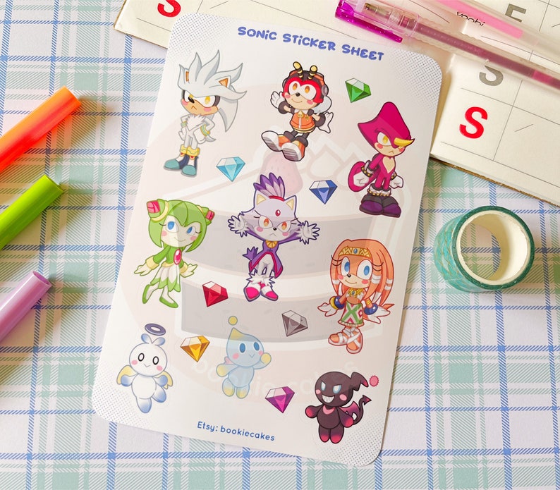 May include: A sticker sheet featuring characters from the Sonic the Hedgehog video game franchise. The sheet includes Sonic, Tails, Knuckles, Amy, Shadow, Rouge, Blaze, and Cream. The characters are depicted in a cartoon style and are surrounded by colorful gems.