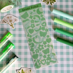 May include: A sheet of green and white holographic stickers with various shapes, including stars, hearts, moons, and butterflies. The stickers are on a green and white checkered background with green and white pens and floral stickers.