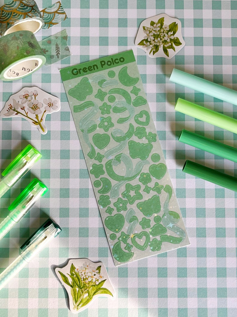 May include: A sheet of green and white holographic stickers with various shapes, including stars, hearts, moons, and butterflies. The stickers are on a green and white checkered background with green and white pens and floral stickers.