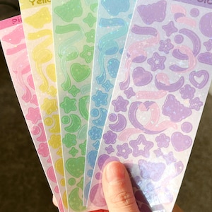 May include: Five sheets of iridescent holographic stickers with various shapes and designs. The stickers are in rainbow colors: pink, yellow, green, blue, and purple. Each sheet has a different color and features a variety of shapes, including stars, hearts, moons, butterflies, and ribbons.