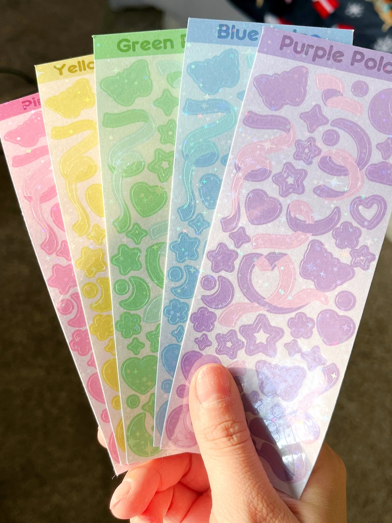 May include: Five sheets of iridescent holographic stickers with various shapes and designs. The stickers are in rainbow colors: pink, yellow, green, blue, and purple. Each sheet has a different color and features a variety of shapes, including stars, hearts, moons, butterflies, and ribbons.