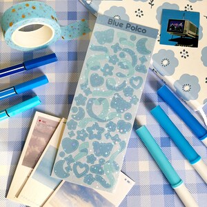 May include: A sheet of blue holographic stickers with a variety of shapes, including stars, hearts, moons, and butterflies. The stickers are on a white background with a blue and white checkered pattern.