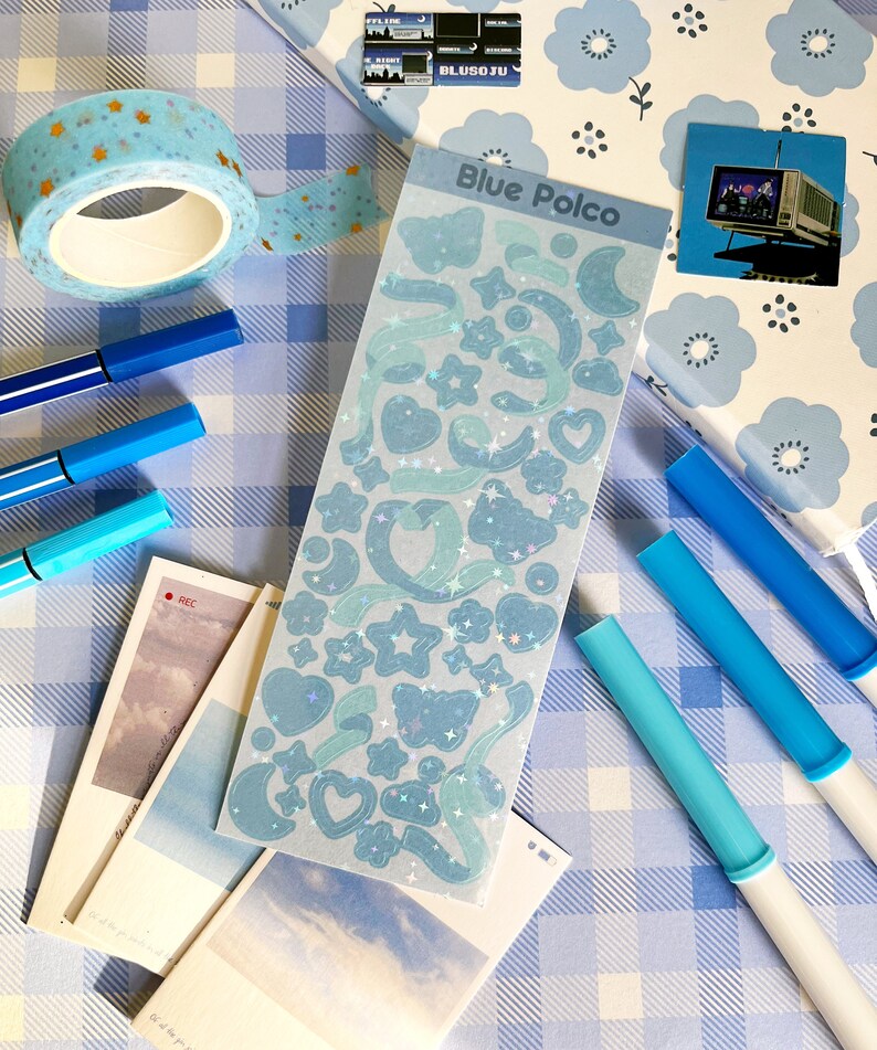 May include: A sheet of blue holographic stickers with a variety of shapes, including stars, hearts, moons, and butterflies. The stickers are on a white background with a blue and white checkered pattern.