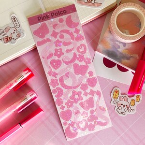 May include: Pink Polco sticker sheet with holographic pink stars, hearts, moons, and butterflies. The sheet is on a pink surface with two pink pens and a pink and white notebook.