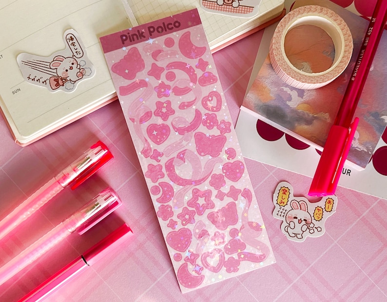 May include: Pink Polco sticker sheet with holographic pink stars, hearts, moons, and butterflies. The sheet is on a pink surface with two pink pens and a pink and white notebook.