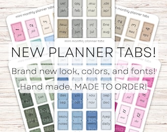May include: A collection of adhesive mini monthly planner tabs arranged on multiple white sticker sheets. The tabs feature various pastel and muted color palettes, including pink, blue, green, and neutral tones, with printed month names and labels.