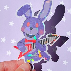 Pu&ograve; includere: Un adesivo olografico viola e blu di un coniglio dei cartoni animati che suona una chitarra elettrica. Il coniglio ha gli occhi rossi e un papillon rosso. La chitarra &egrave; rossa con corde nere.