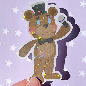 Pu&ograve; includere: Un adesivo olografico di un orso marrone che indossa un cappello a cilindro nero e un papillon, tenendo un microfono. L'orso ha gli occhi blu e una texture scintillante.