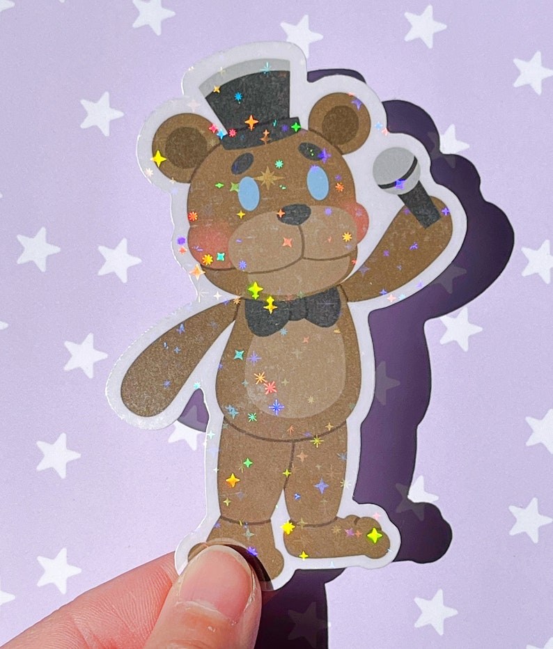 Pu&ograve; includere: Un adesivo olografico di un orso marrone che indossa un cappello a cilindro nero e un papillon, tenendo un microfono. L'orso ha gli occhi blu e una texture scintillante.