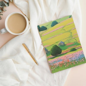 May include: A white notebook with a green, yellow, and pink landscape illustration on the cover. The notebook is open and lying on a white blanket with a cup of coffee and a gold pen.