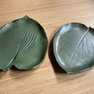 May include: Two leaf-shaped decorative trays. The trays are a muted green color with detailed veining, and are made of a metallic material. The trays are placed on a wooden surface.