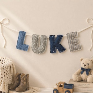 May include: A decorative banner spelling "LUKE" in fabric letters, each with a unique pattern and color, suspended by a white cord. The scene includes a teddy bear, books, a toy truck, and a pair of cowboy boots, suggesting a nursery theme.