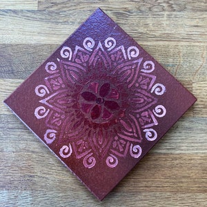 May include: A square canvas painting featuring a pink and red mandala design. The mandala is painted in a gradient of pink and red, with a darker red centre. The mandala is surrounded by a white border.