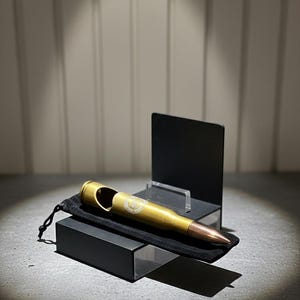 Puede incluir: Un abridor de botellas con forma de bala, color latón, con punta cobriza. Descansa sobre una bolsa de terciopelo negro dentro de una caja negra, con un expositor negro detrás. El abridor tiene una abertura circular.