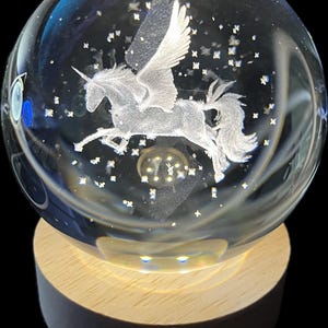 May include: A clear glass sphere with a white, winged unicorn etched inside. The sphere sits on a wooden base with a light shining from the bottom.