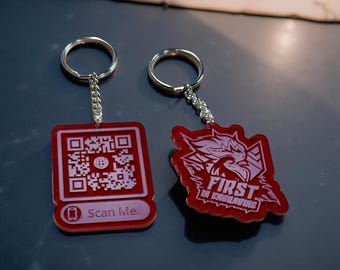 Puede incluir: Dos llaveros rojos de acrílico con anillas plateadas. Uno es cuadrado con un código QR y el texto "Scan Me". El otro es un diseño estilizado con el texto "FIRST IN ENGRAVING".