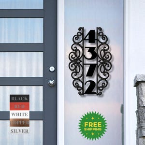 May include: Black metal house number plaque with a decorative scroll design. The numbers 4372 are displayed vertically. The plaque is mounted on a white door.
