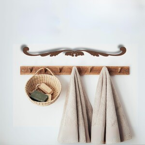 May include: A wooden wall-mounted rack with hooks for hanging towels. Two towels are hanging on the hooks. A wicker basket with soap and brushes is hanging on the left hook. A decorative wooden scroll is mounted above the rack.