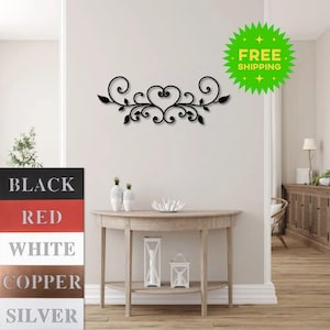 May include: Black metal wall decor with a heart and scroll design. The decor is shown hanging on a white wall above a wooden console table. The image also shows a variety of color options for the decor: black, red, white, copper, and silver.