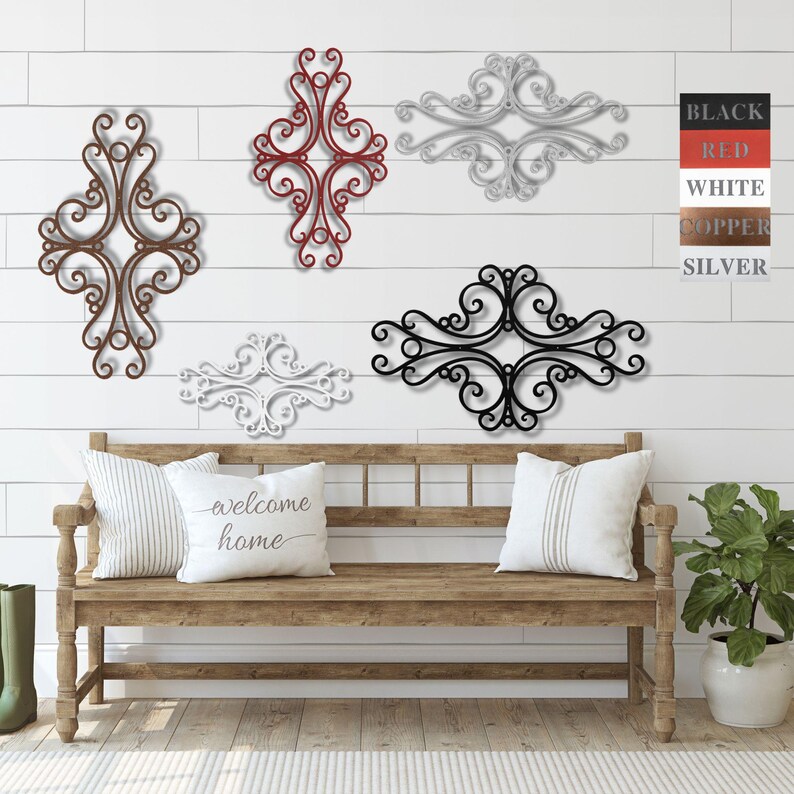 May include: Five decorative metal wall decor pieces in various styles and sizes. The pieces are in black, white, red, copper, and silver. The decor is displayed on a white wall with a wooden bench in front of it.