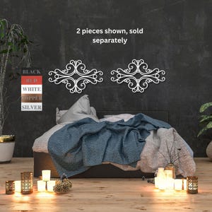 May include: Two white metal wall decor pieces with a decorative scroll design. The pieces are shown on a black wall above a bed with a blue and white blanket. The text "2 pieces shown, sold separately" is displayed above the decor.