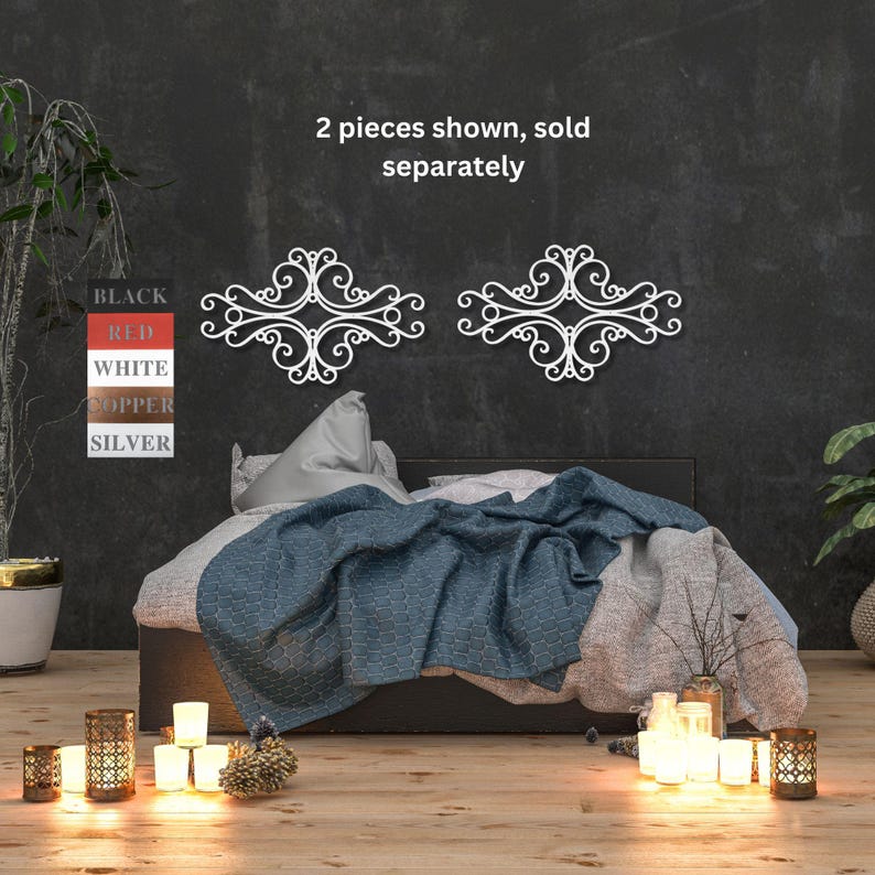 May include: Two white metal wall decor pieces with a decorative scroll design. The pieces are shown on a black wall above a bed with a blue and white blanket. The text "2 pieces shown, sold separately" is displayed above the decor.