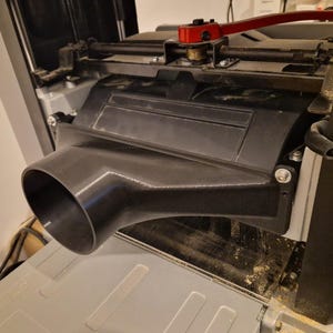 Può includere: Primo piano di una macchina per la lavorazione del legno grigia e nera con una porta di aspirazione della polvere nera. La macchina ha una maniglia rossa ed è coperta di segatura. La macchina viene utilizzata per la lavorazione del legno.