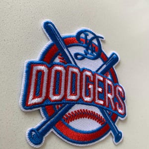 May include: An embroidered patch featuring the word DODGERS in red letters with a blue outline. The design includes a white baseball with red stitching, a red circular border, and two crossed blue baseball bats. A stylized blue letter D sits above.