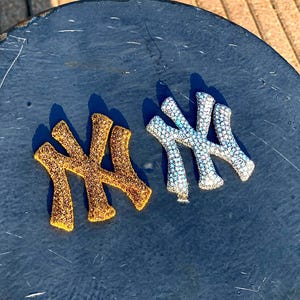 May include: Two New York Yankees logo patches, one covered in gold-toned rhinestones and the other in silver-toned rhinestones. Each patch features the iconic interlocking NY letters, with a textured, sparkling surface created by the dense arrangement of small, reflective stones.