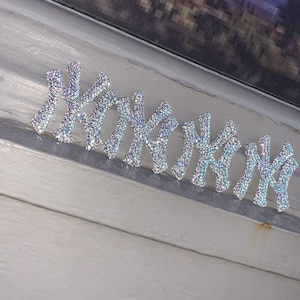 May include: A row of four silver-toned New York Yankees logo emblems covered in iridescent, light-reflecting rhinestones. Each emblem features the iconic interlocking NY letters, creating a sparkling, multi-colored effect under the light.