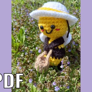 May include: A crocheted bee toy with a yellow body, black stripes, and a white hat. The bee is holding a brown basket and has a smiling face with black eyes and pink cheeks.