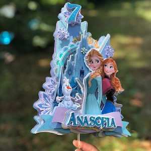 May include: A multi-layered cake topper featuring Elsa and Anna from Frozen, with a castle backdrop and the number 7. The topper is in shades of blue, purple, and white, with the name "ANASOFIA" displayed on a banner.
