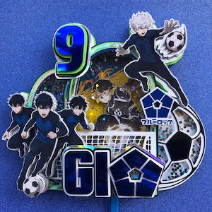 May include: A blue and green cake topper with a number 9, a soccer goal, a soccer ball, and characters from the anime "Blue Lock". The text "GK" is on the cake topper.