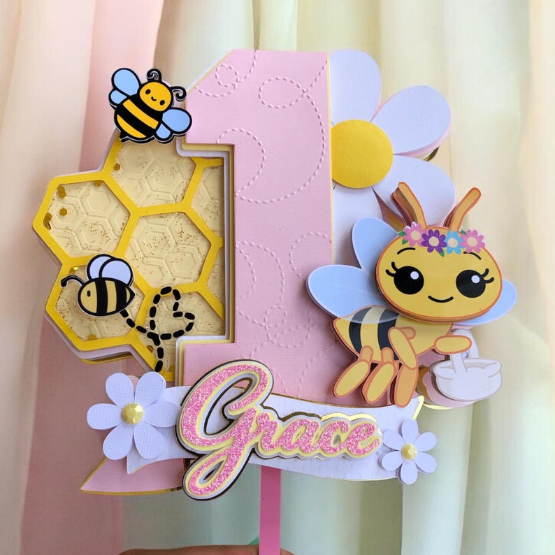 May include: A pink and yellow number one cake topper with a bee theme. The topper features a honeycomb, bees, flowers, and the name "Grace" in glittery pink. The design is layered and three-dimensional, perfect for a first birthday celebration.
