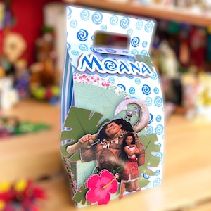 May include: A blue and green party favor box with a cutout handle. The box features a cartoon image of Moana and Maui from the Disney movie Moana. The box has the word "Moana" printed on the front.