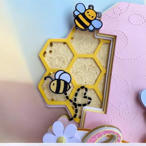 May include: A layered paper craft featuring a yellow honeycomb design with glitter accents and two cartoon bees. The background is a gradient of pink, yellow, and blue. A white flower and a rainbow are visible at the bottom.
