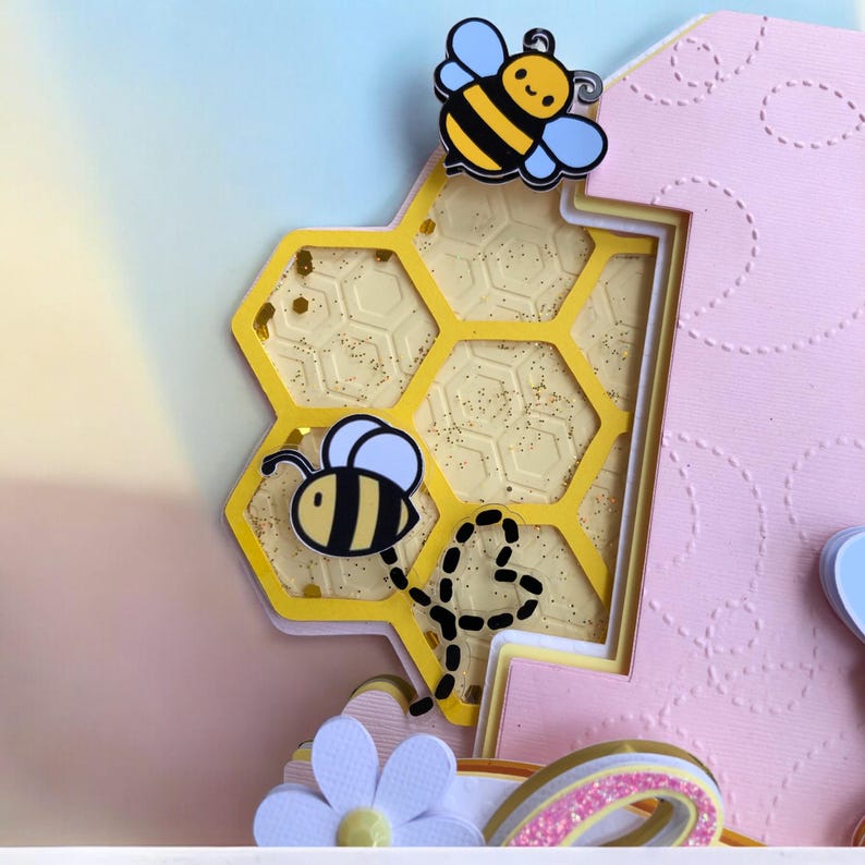 May include: A layered paper craft featuring a yellow honeycomb design with glitter accents and two cartoon bees. The background is a gradient of pink, yellow, and blue. A white flower and a rainbow are visible at the bottom.