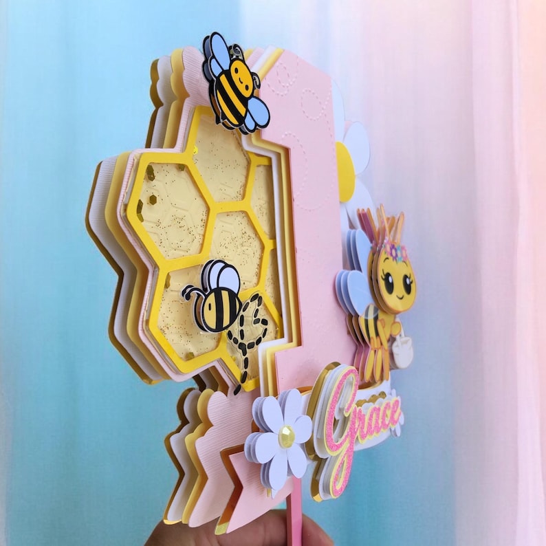 May include: A pink and yellow layered number one cake topper with bee and honeycomb details. The topper features a cartoon bee, a honeycomb design, and the name "Grace" in cursive. A white flower and blue accents complete the design.