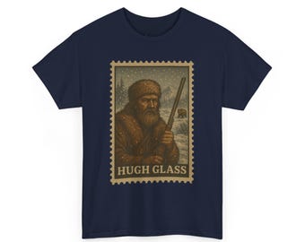 May include: Navy blue t-shirt featuring a vintage postage stamp design with the text "HUGH GLASS". The stamp depicts a man in a fur hat and coat holding a rifle in a snowy landscape with a bear in the background.