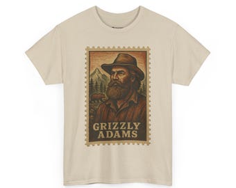 May include: Beige t-shirt featuring a vintage-style Grizzly Adams stamp design. The stamp depicts a portrait of a man with a beard and hat, with a bear and mountain scene in the background. The words "GRIZZLY ADAMS" are printed at the bottom.