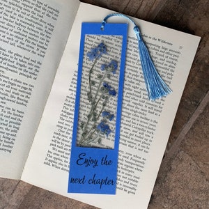 Könnte beinhalten: Blaues Lesezeichen mit einem gepressten Blumenmuster und dem Text "Enjoy the next chapter".