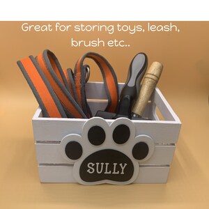 May include: White wooden crate with a paw print design and the name "SULLY" on it. The crate is filled with a leash, a dog brush, and other dog accessories.