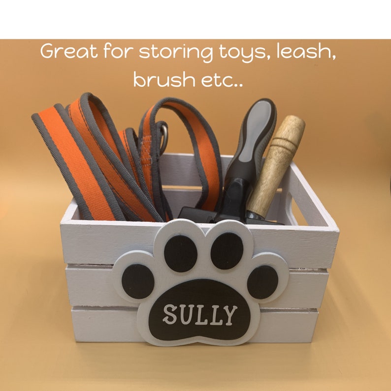 May include: White wooden crate with a paw print design and the name "SULLY" on it. The crate is filled with a leash, a dog brush, and other dog accessories.