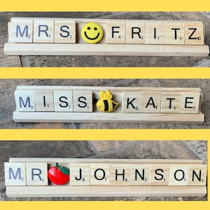 Three wooden nameplates with letter tiles spelling out 'MRS FRITZ', 'MISS KATE', and 'MR JOHNSON'. The nameplates are decorated with a smiley face, a bee, and an apple, respectively.
