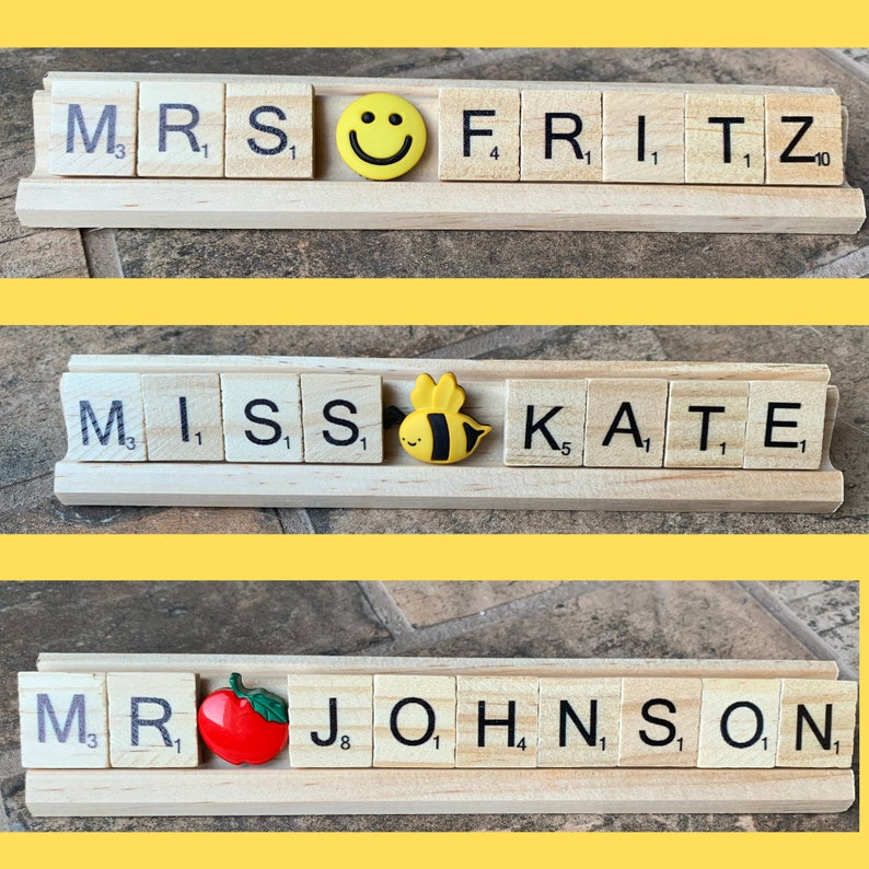Three wooden nameplates with letter tiles spelling out 'MRS FRITZ', 'MISS KATE', and 'MR JOHNSON'. The nameplates are decorated with a smiley face, a bee, and an apple, respectively.