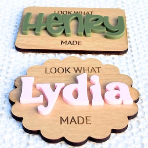 May include: Two wooden name plaques. One is rectangular with the name "Henry" in green letters. The other is scalloped with the name "Lydia" in pink letters. Both plaques have the text "LOOK WHAT MADE" engraved.